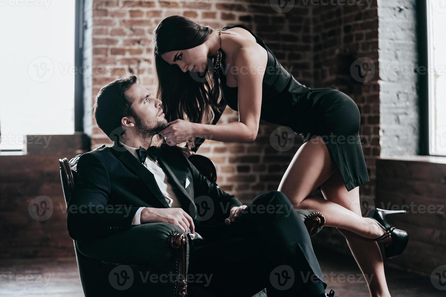 tempo per sedurre. bellissimo giovane donna nel cocktail vestito pendente per sua fidanzato seduta nel sedia mentre guardare a ogni altro nel soffitta interno foto