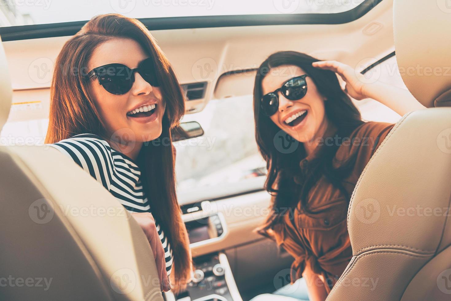 pronto per strada viaggio posteriore Visualizza di Due bellissimo giovane allegro donne guardare a telecamera con Sorridi mentre seduta nel auto foto