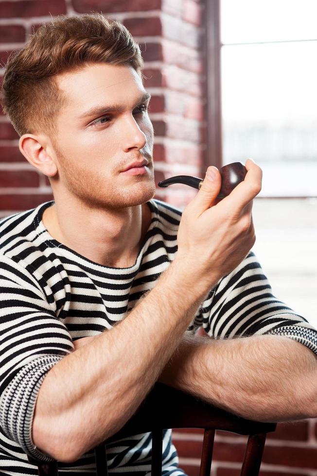 fumo tubo. bello giovane uomo nel a strisce camicia fumo tubo e guardare lontano mentre seduta su il sedia foto