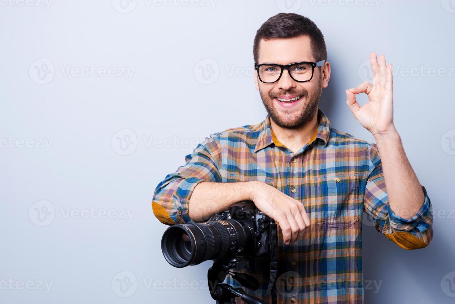 amore mio lavoro. ritratto di fiducioso giovane uomo nel camicia Tenere mano su telecamera su tripode e Esprimere a gesti mentre in piedi contro grigio sfondo foto