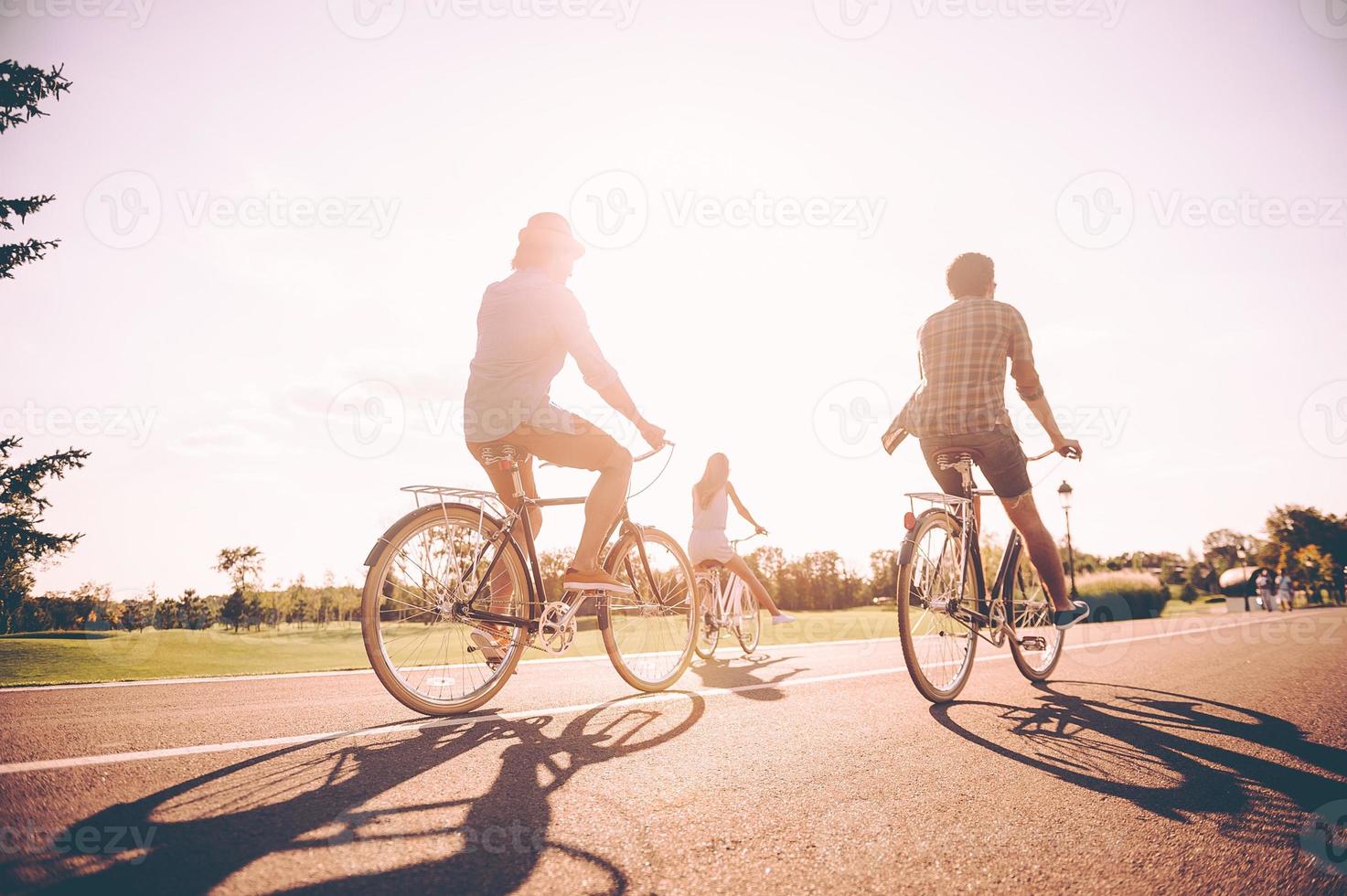 godendo spensierato tempo insieme. posteriore Visualizza di giovane allegro persone equitazione biciclette lungo un' strada insieme foto