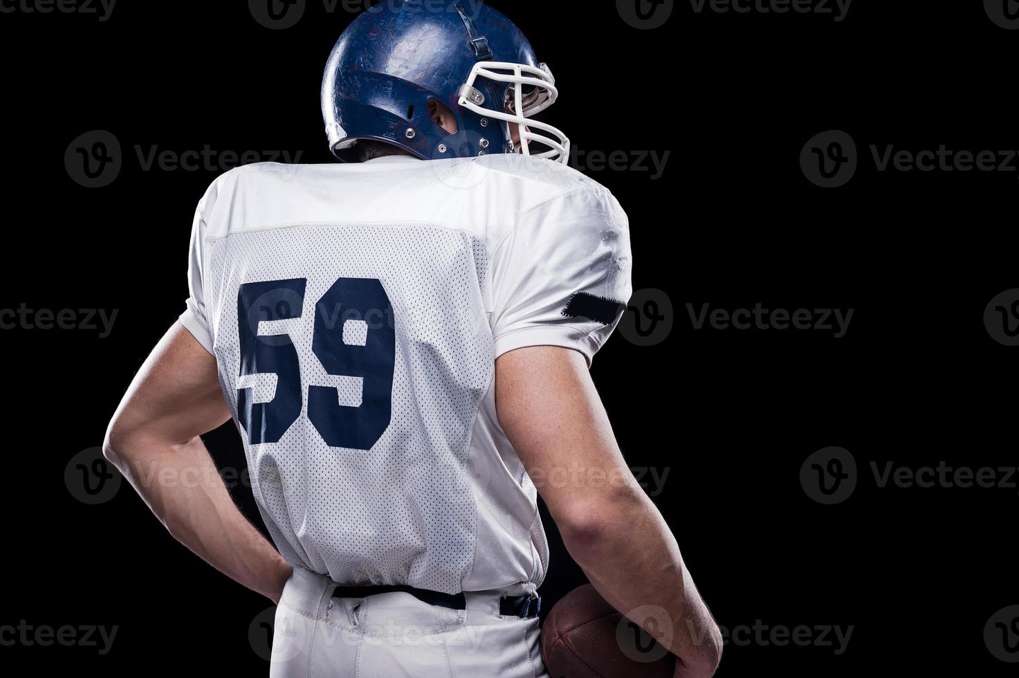 potente Indietro. posteriore Visualizza di americano calcio giocatore Tenere calcio palla mentre in piedi contro nero sfondo foto