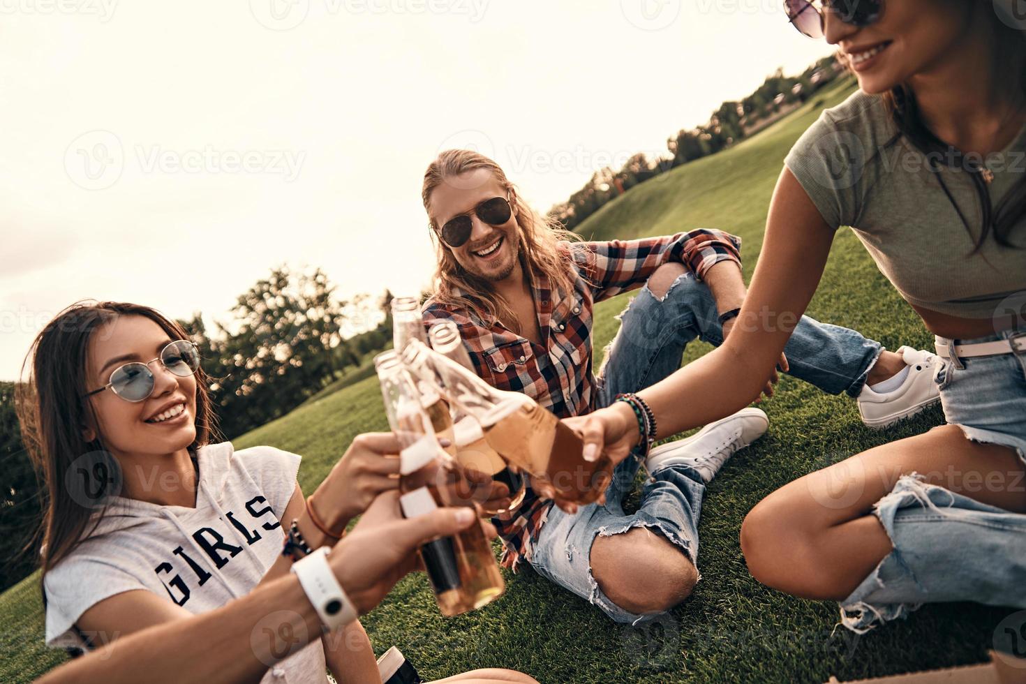 gruppo di giovane sorridente persone nel casuale indossare tostatura con birra bottiglie mentre godendo picnic all'aperto foto