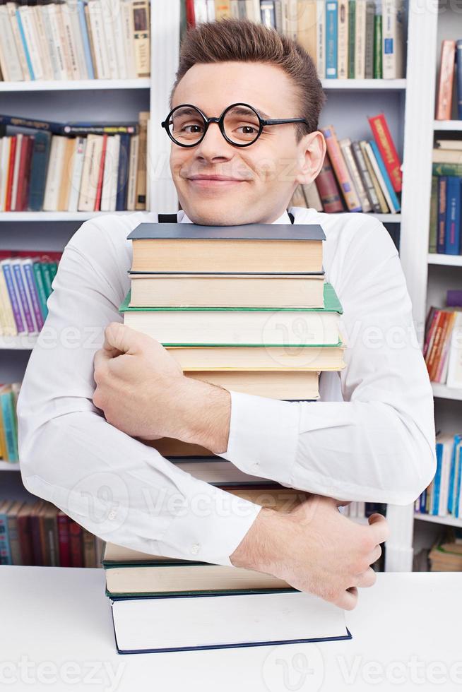 io amore studiando. allegro giovane nerd uomo nel camicia e arco cravatta seduta a il tavolo nel biblioteca e abbracciare un' libro pila foto