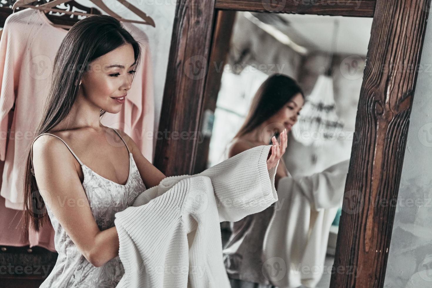 Perfetto camicetta. attraente giovane donna assunzione su il camicetta e sorridente mentre in piedi nel davanti di il specchio a casa foto