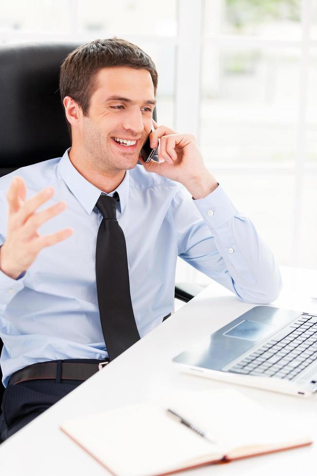 contento uomo d'affari. contento giovane uomo nel vestito formale guardare a computer tenere sotto controllo e parlando su il mobile Telefono mentre seduta a il suo Lavorando posto foto