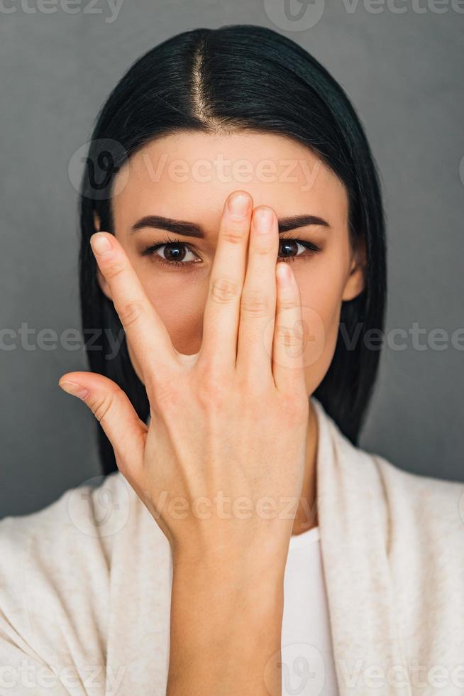 nascondere e cercare. ritratto di bellissimo giovane donna guardare a telecamera attraverso sua dita mentre in piedi contro grigio sfondo foto