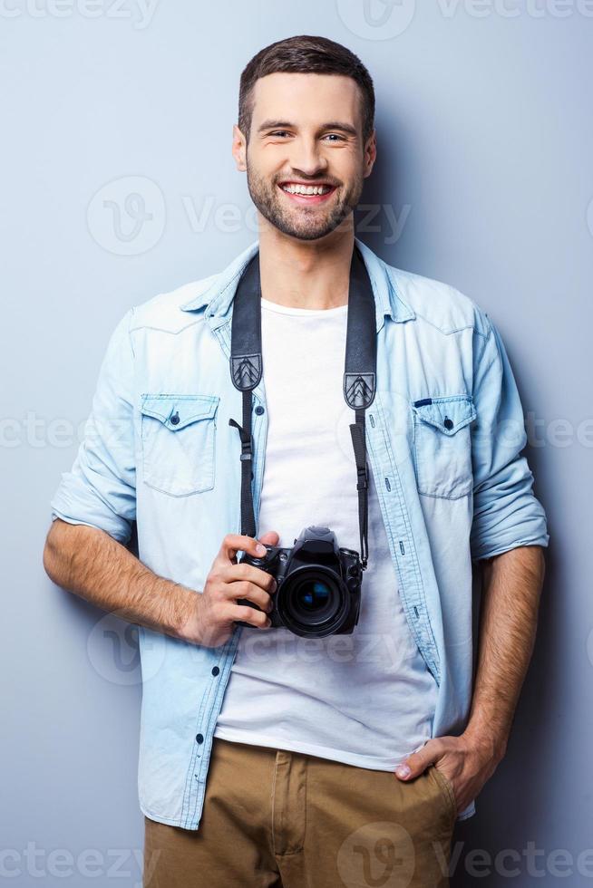 fiducioso fotografo. bello giovane uomo Tenere digitale telecamera e sorridente mentre in piedi contro grigio sfondo foto