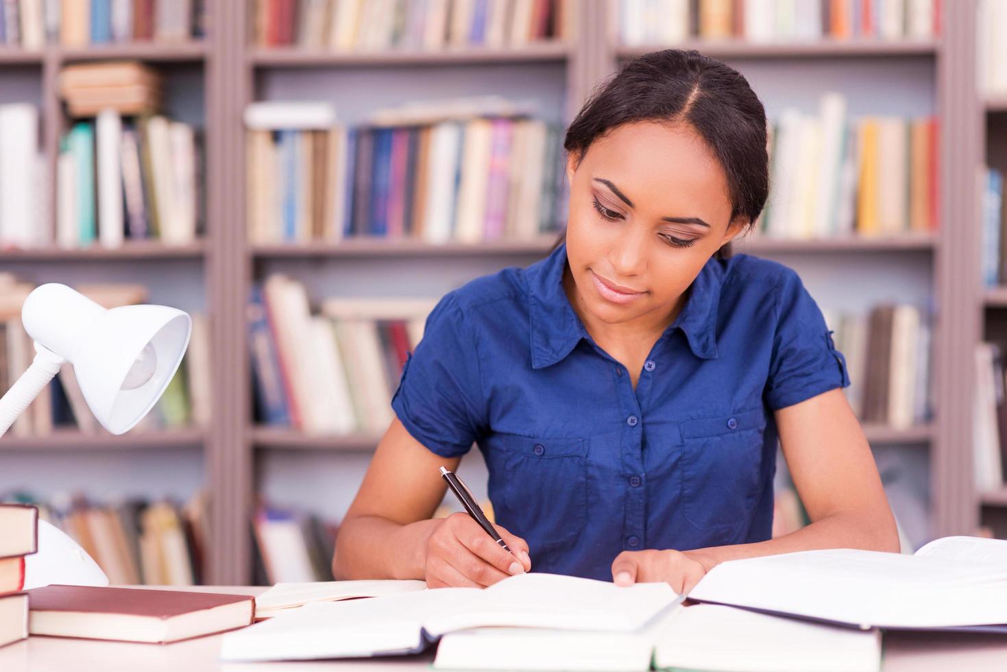 alunno preparazione per il esami. fiducioso giovane nero donna scrittura qualcosa nel sua Nota tampone e lettura libro mentre seduta a il biblioteca scrivania foto