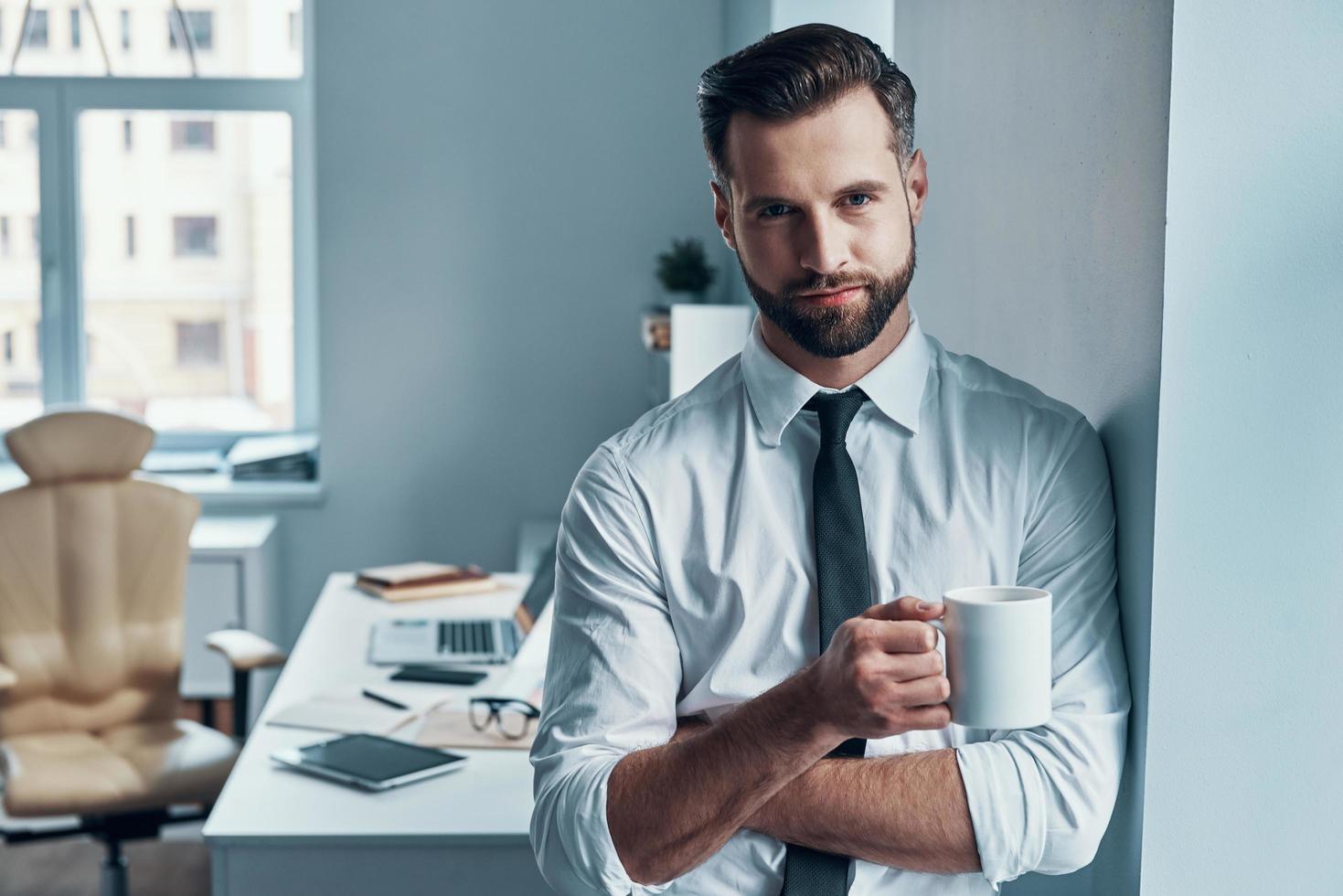 bene guardare giovane uomo nel camicia e cravatta Tenere tazza e sorridente mentre in piedi nel il ufficio foto