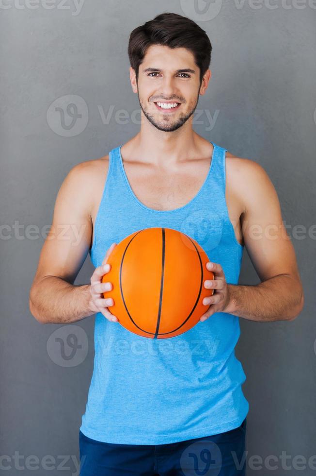 io amore pallacanestro sorridente giovane muscolare uomo Tenere pallacanestro palla mentre in piedi contro grigio sfondi foto