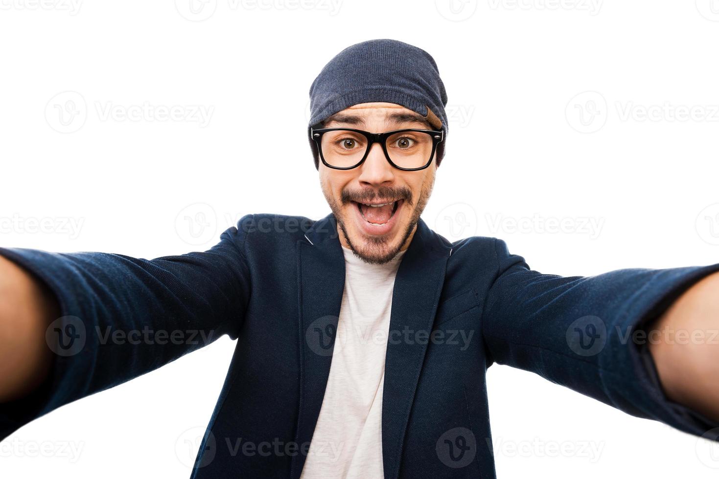 appena me allegro giovane uomo nel occhiali e cappello fabbricazione autoscatto mentre in piedi contro bianca sfondo foto