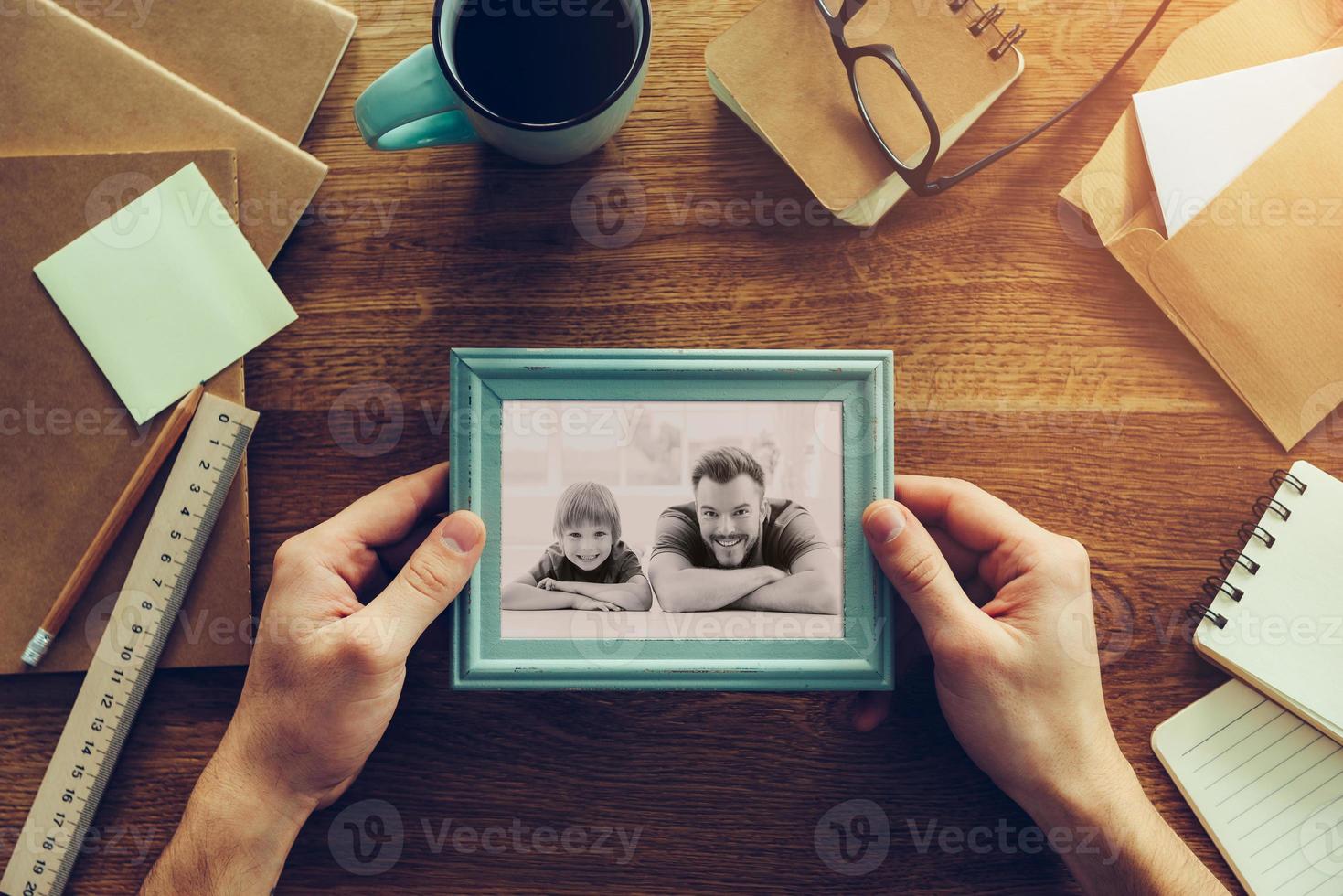 mio figlio è mio ispirazione. avvicinamento superiore Visualizza di uomo Tenere fotografia di lui stesso e il suo figlio al di sopra di di legno scrivania con diverso cancelleria cose posa in giro foto