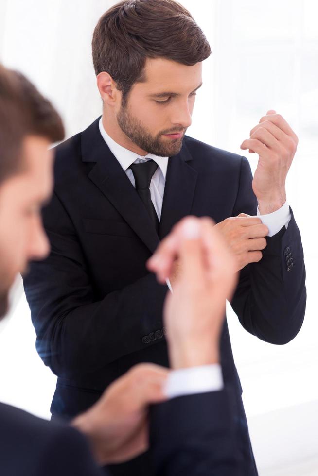 qualunque cosa dovrebbero essere Perfetto. bello giovane uomo nel vestito formale regolazione il suo maniche mentre in piedi contro specchio foto