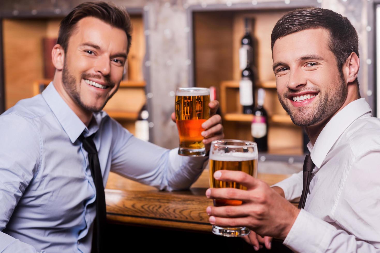 rilassante dopo difficile Lavorando giorno. Due contento giovane uomini nel camicia e cravatta Tenere bicchieri con birra e sorridente mentre seduta a il bar contatore foto