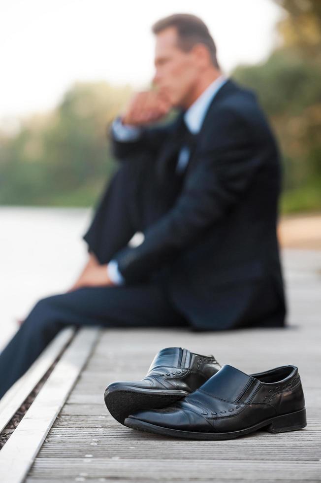 lui esigenze per restare solo adesso. lato Visualizza di riflessivo uomo d'affari Tenere mano su mento e guardare lontano mentre seduta a piedi nudi a il banchina e con scarpe posa su primo piano foto