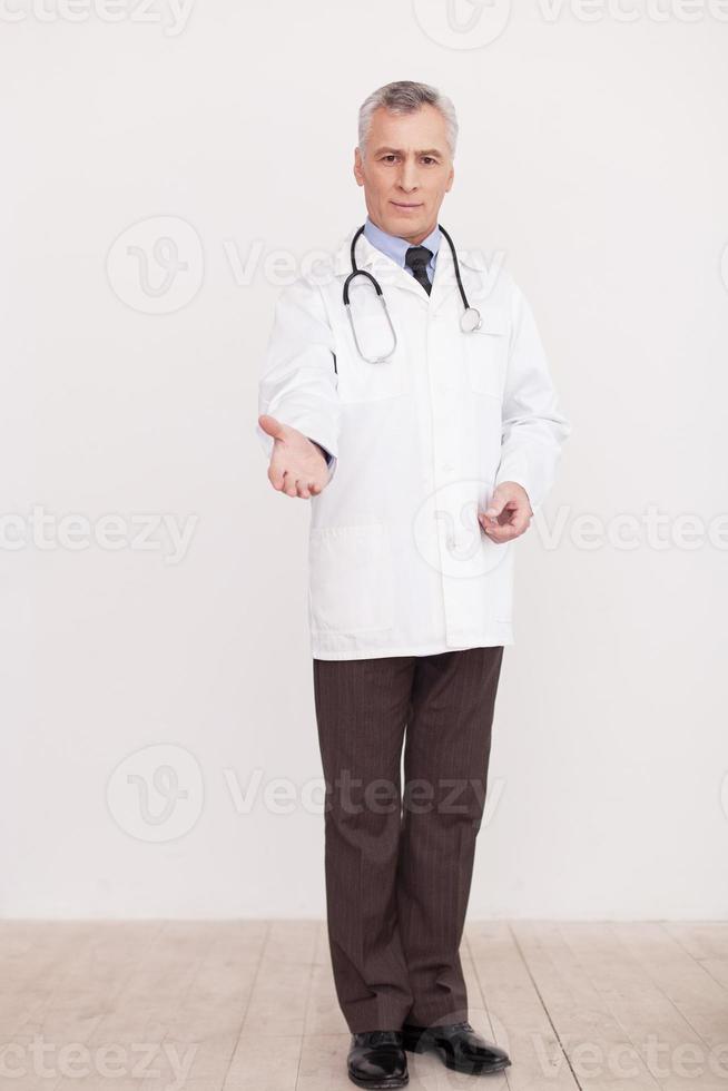 fiducia me pieno lunghezza di anziano grigio capelli medico nel bianca uniforme guardare a telecamera e allungamento su mano mentre in piedi isolato su bianca foto