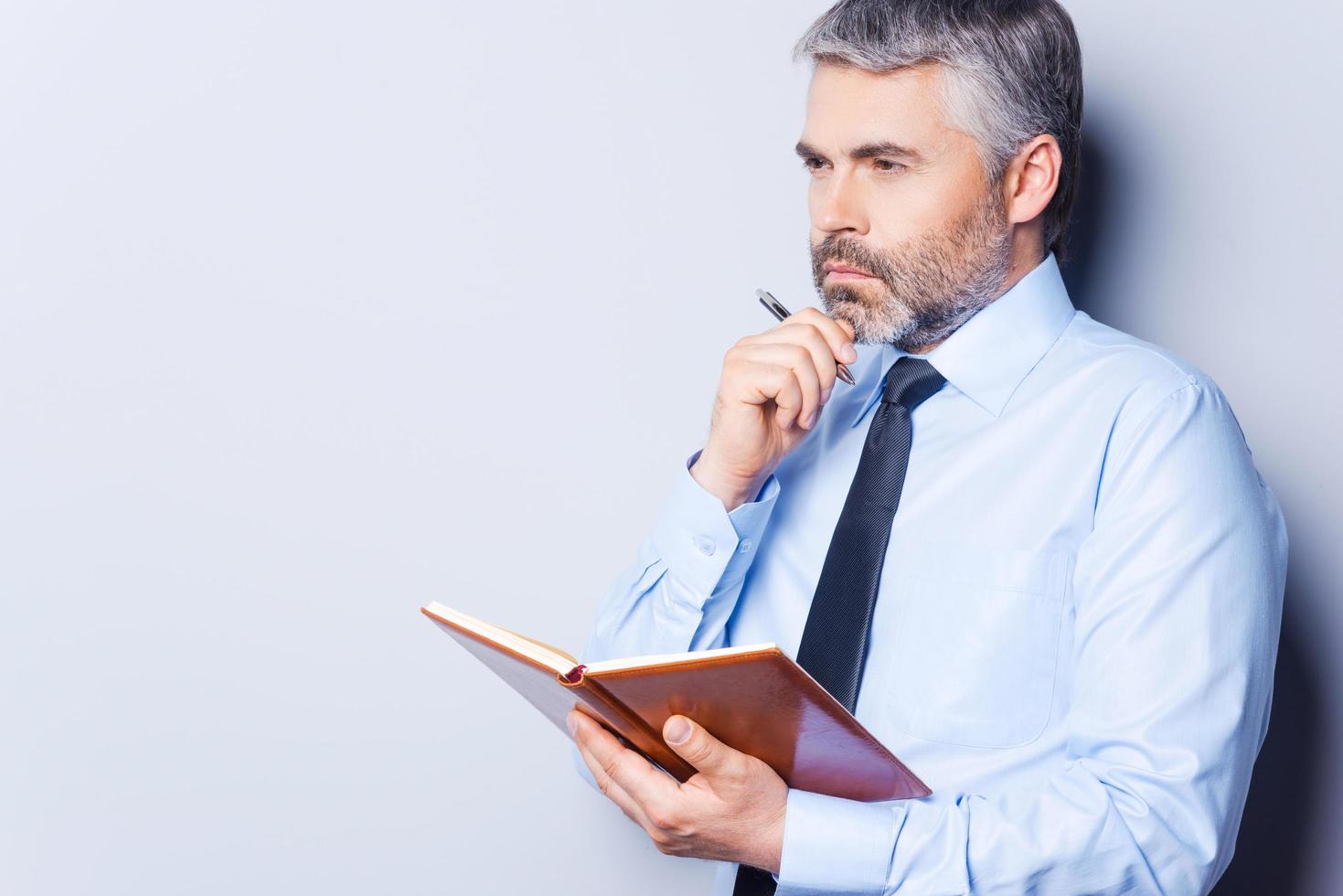 in attesa per ispirazione. riflessivo maturo uomo nel camicia e cravatta toccante il suo mento con mano e guardare lontano mentre Tenere Nota tampone e in piedi contro grigio sfondo foto