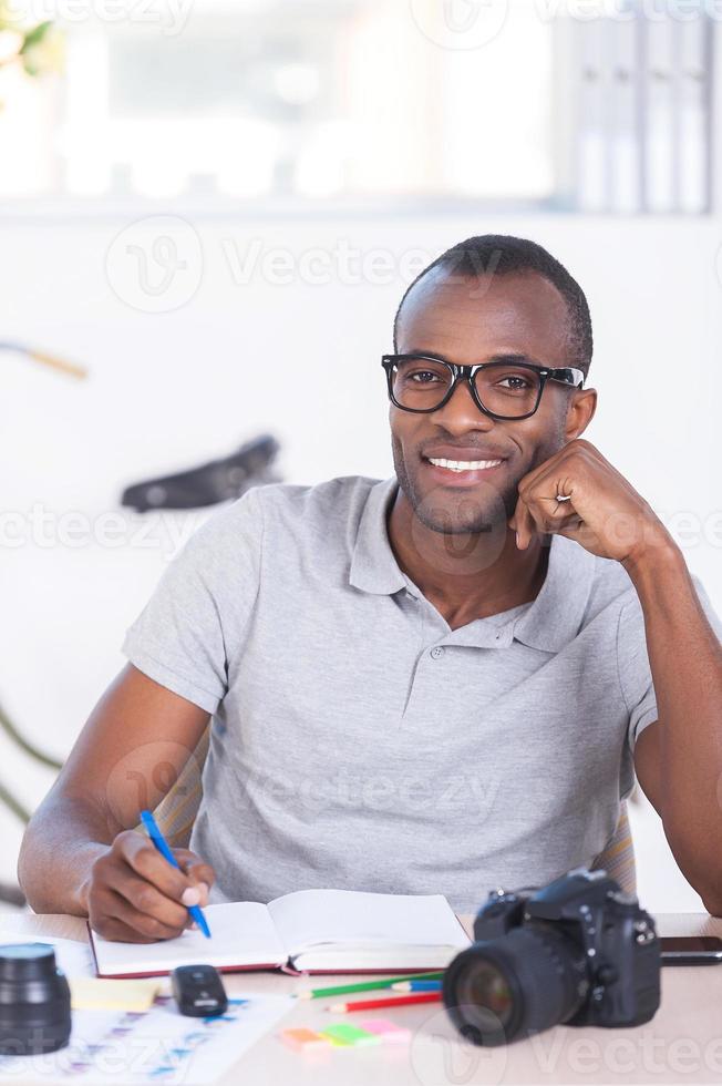 io amore mio lavoro allegro giovane africano uomo nel casuale indossare scrittura qualcosa nel Nota tampone e guardare a telecamera mentre seduta a il suo Lavorando posto foto