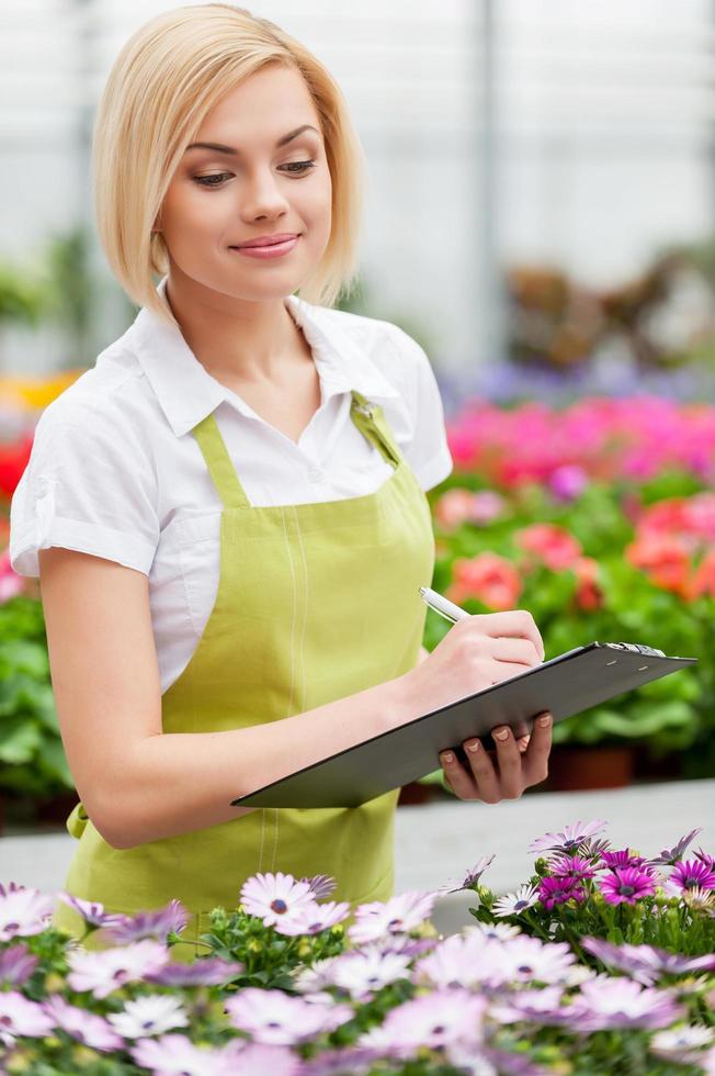 Lavorando nel serra. bellissimo biondo capelli donna nel uniforme scrittura qualcosa nel sua Nota tampone e guardare a il fiori mentre in piedi nel serra foto