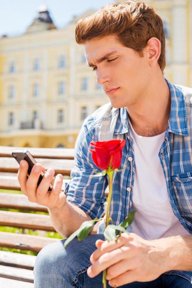 lei è non In arrivo. depresso giovane uomo Tenere singolo rosa e guardare a il suo mobile Telefono mentre seduta su il panchina foto