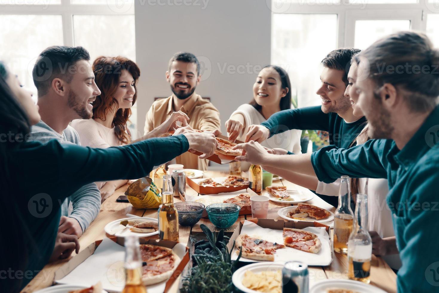 fretta su per raccogliere il migliore fetta gruppo di giovane persone nel casuale indossare raccolta Pizza e sorridente mentre avendo un' cena festa in casa foto