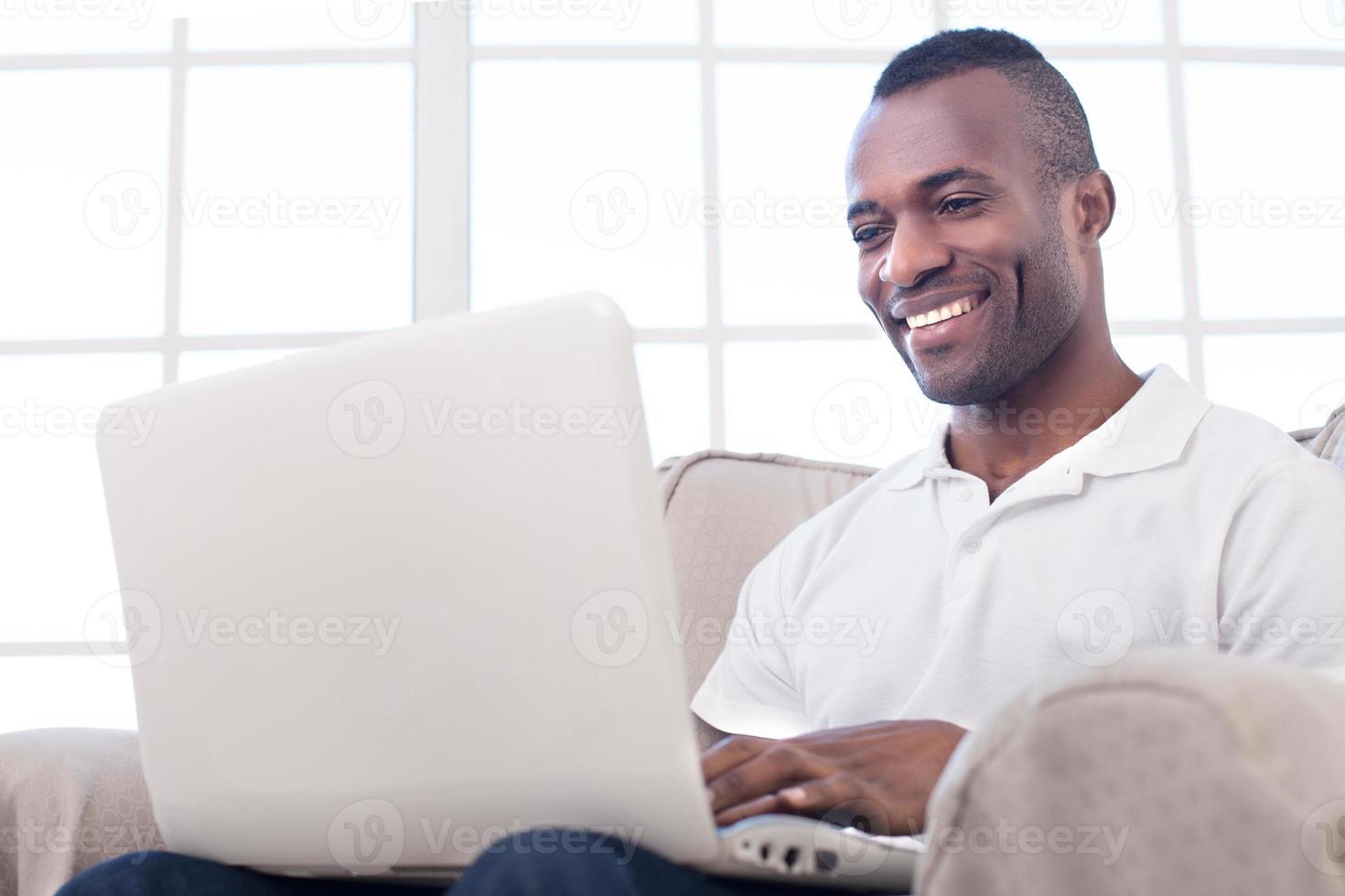 Lavorando a casa. allegro africano uomo utilizzando computer e sorridente mentre seduta su il sedia foto