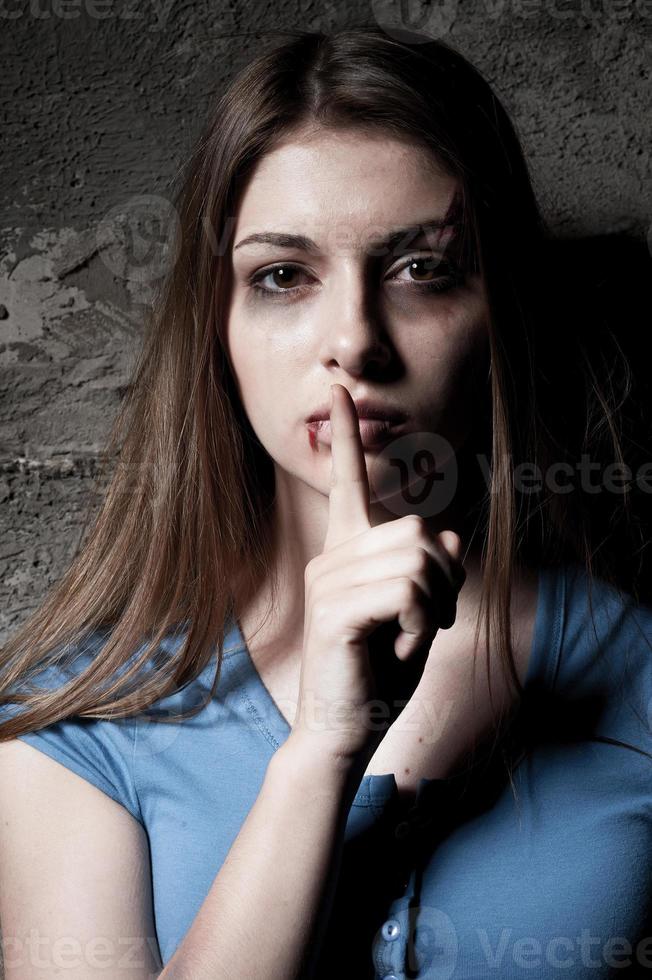 fare non essere silenzioso giovane picchiato su donna guardare a telecamera e Tenere dito su bocca mentre in piedi contro buio parete foto