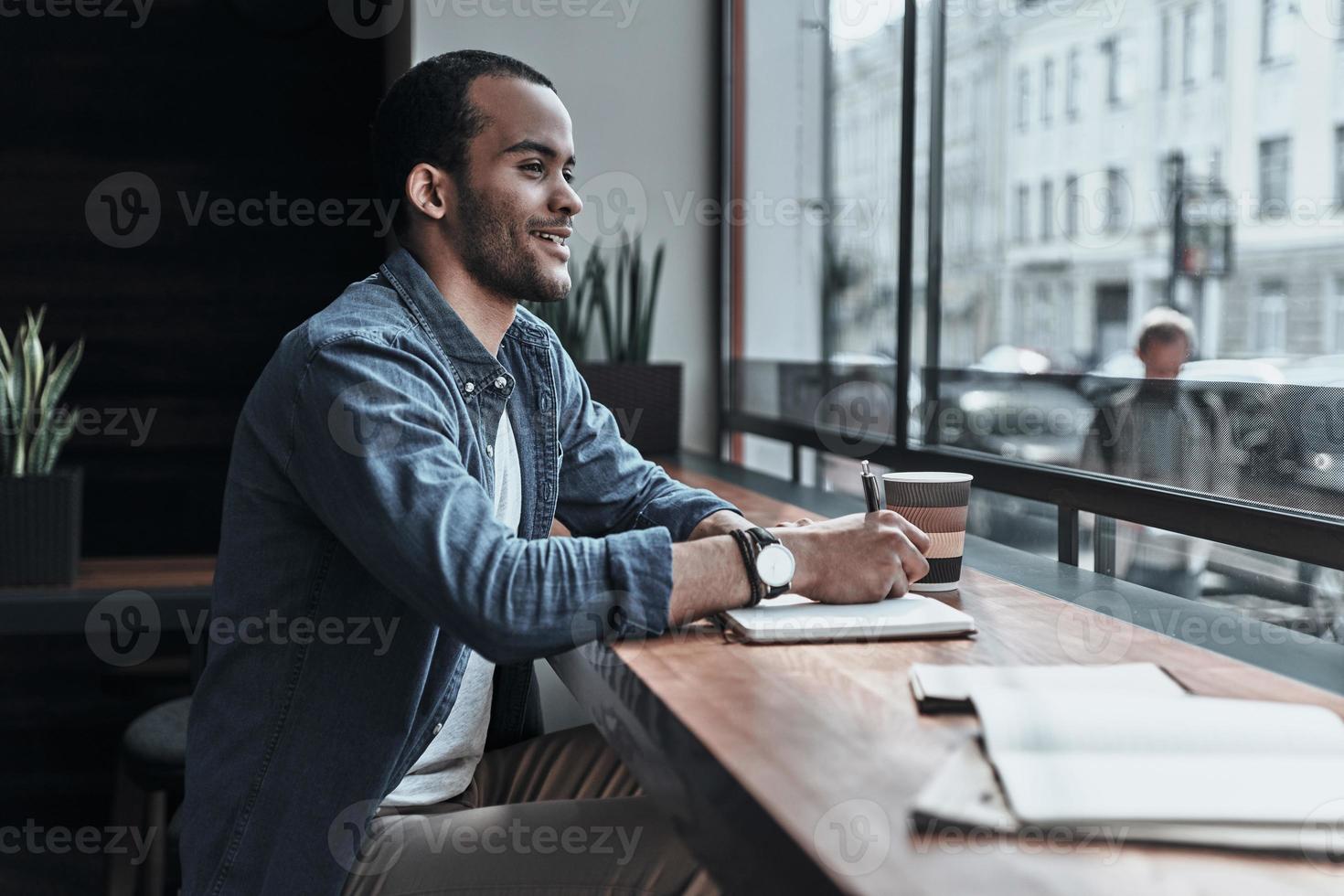 dando il Uscita per il suo idee. bello giovane uomo guardare attraverso il finestra e scrittura qualcosa mentre seduta nel il caffetteria foto