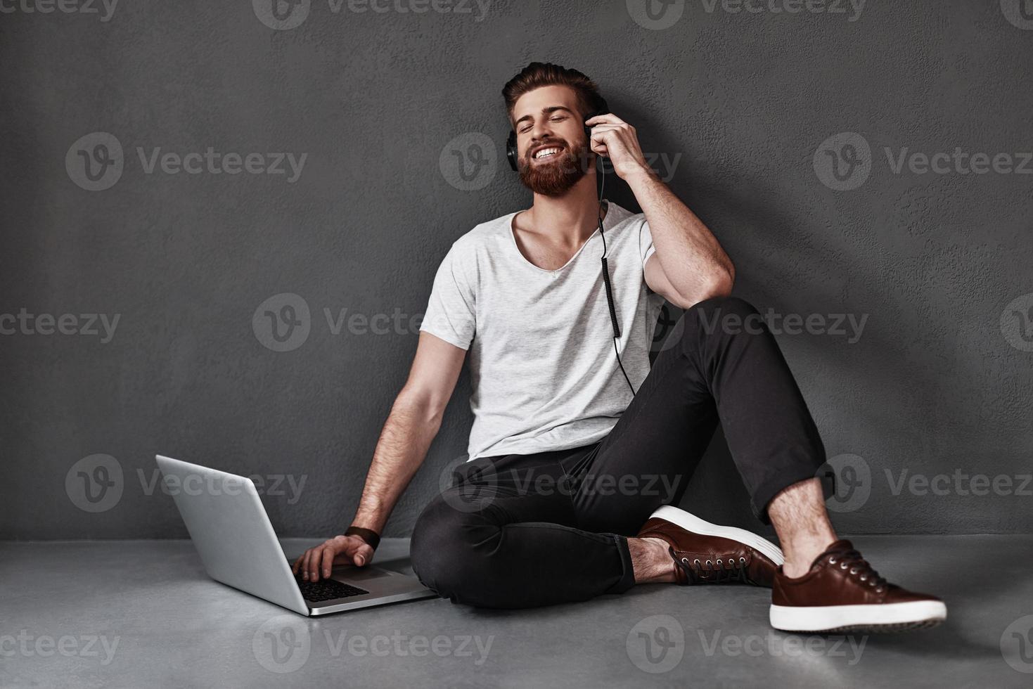 godendo musica. bello giovane uomo ascoltando musica e utilizzando computer mentre seduta su il pavimento contro grigio sfondo foto