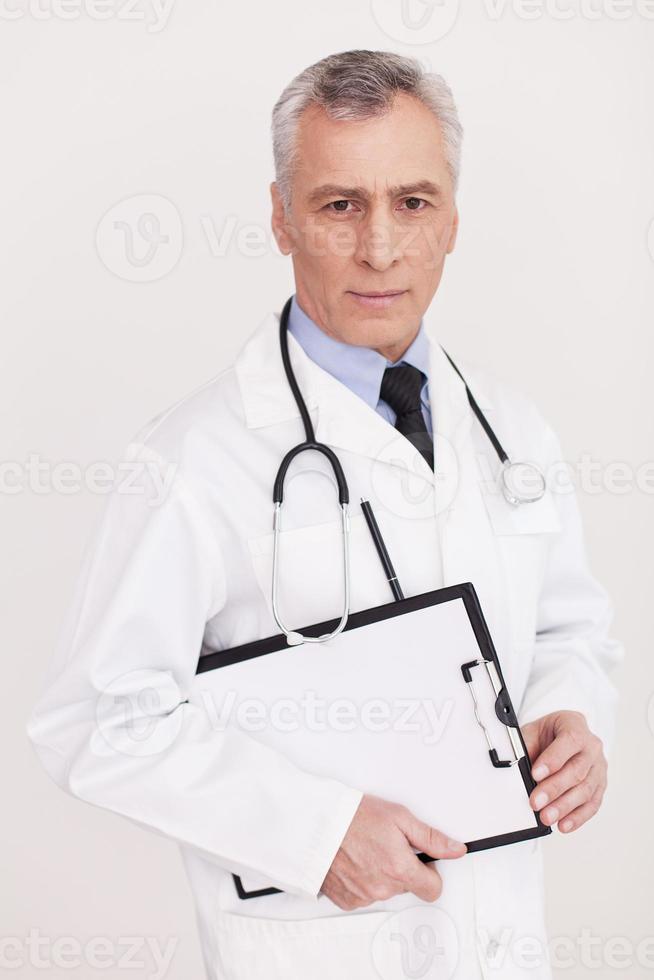 fiducioso medico. anziano grigio capelli medico nel uniforme guardare a telecamera e Tenere un' appunti nel il suo mani mentre in piedi isolato su bianca foto