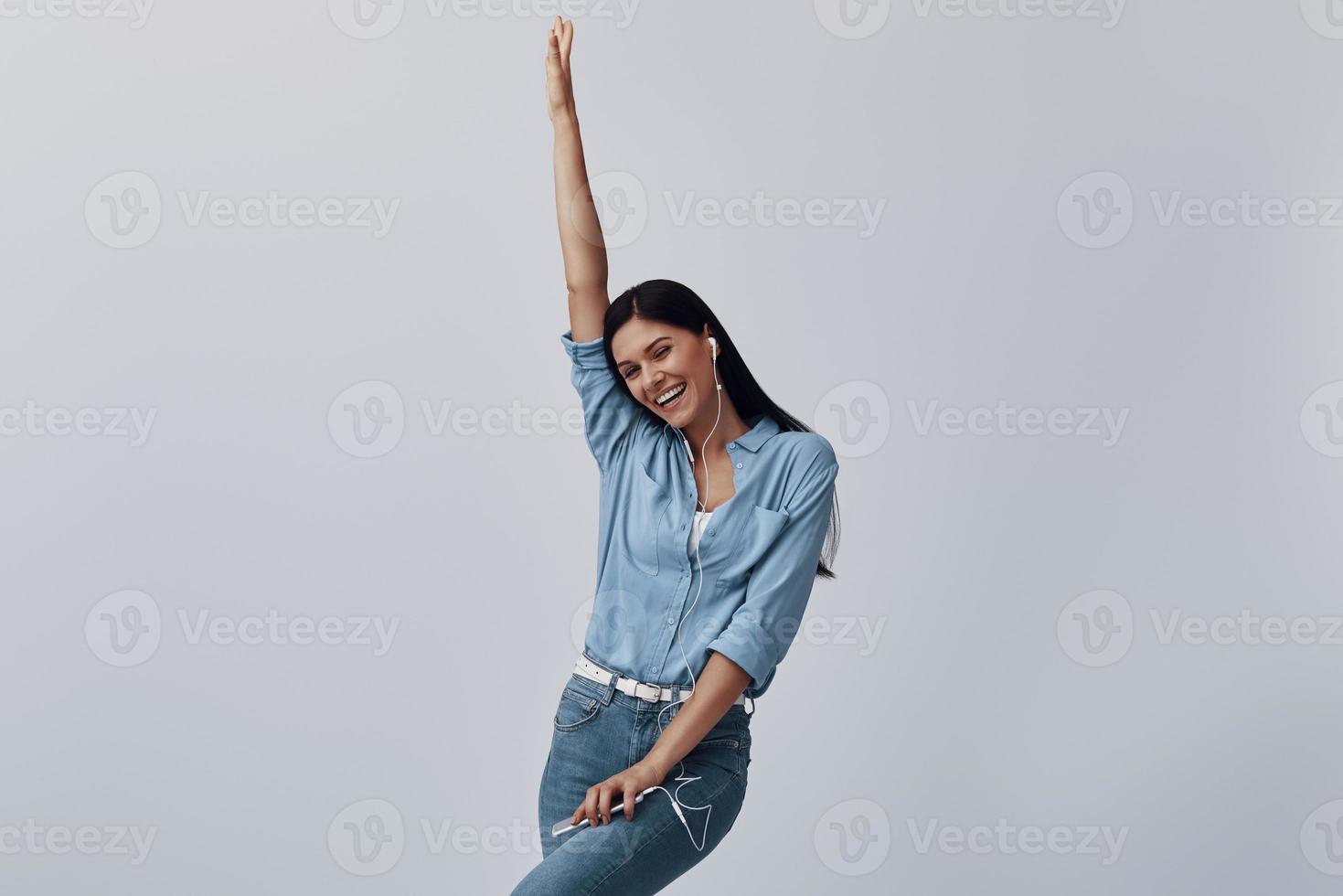 attraente giovane donna ascoltando musica e sorridente mentre in piedi contro grigio sfondo foto