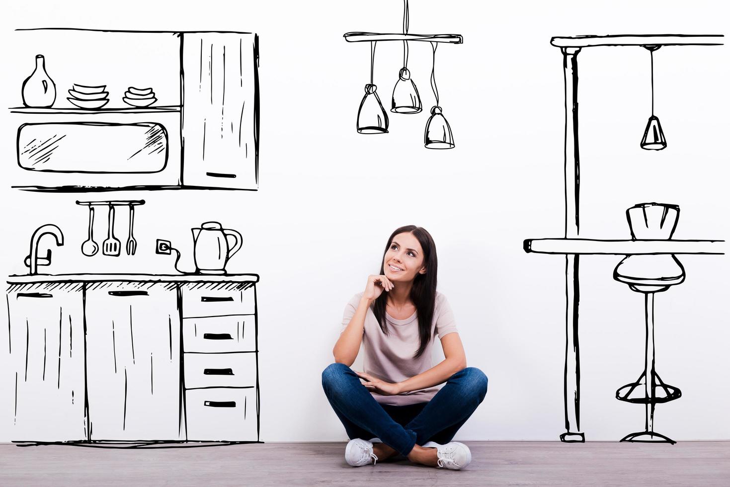 sognare di nuovo cucina. allegro giovane donna sorridente mentre seduta su il pavimento contro bianca sfondo con disegnato cucina foto