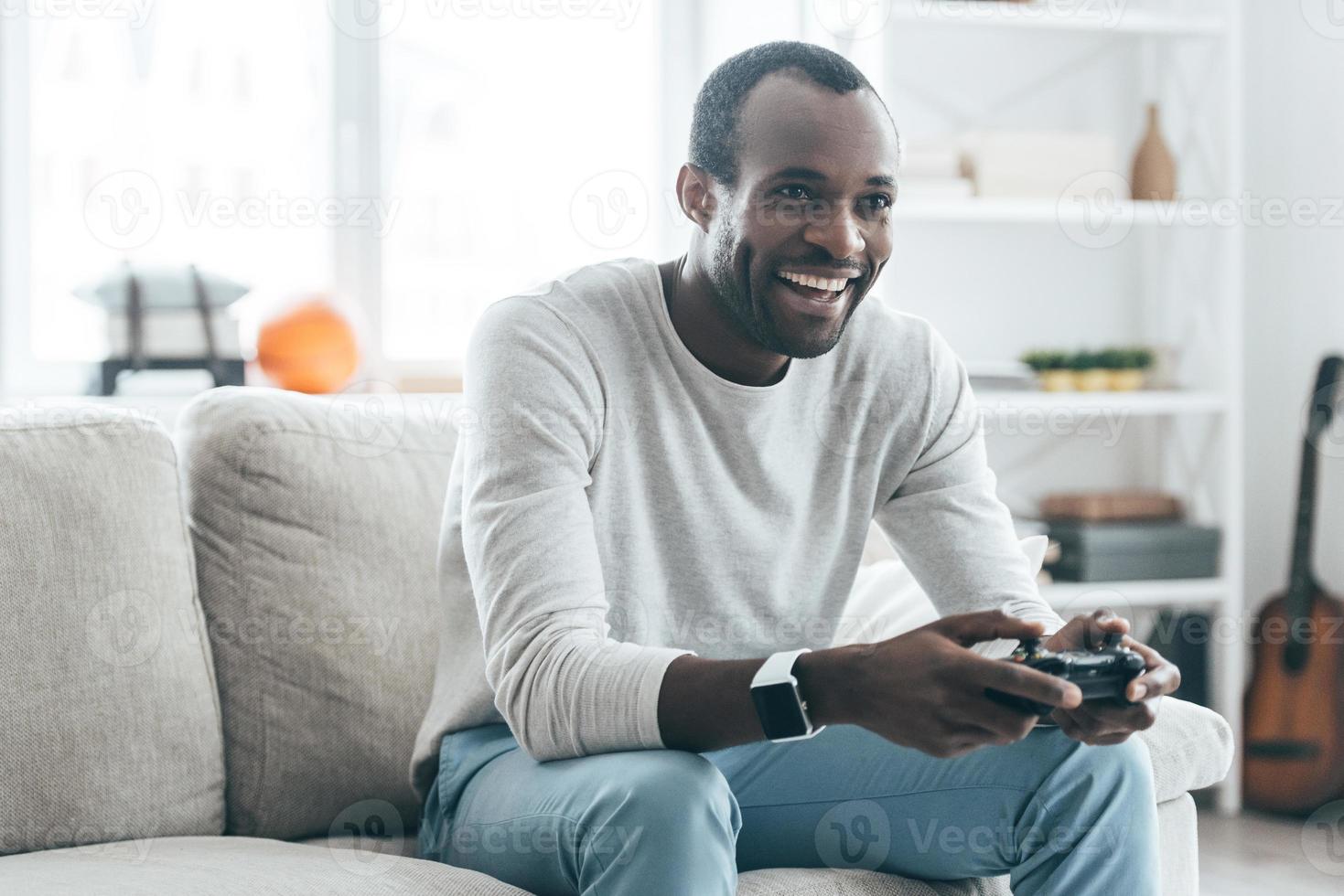 riposo a casa. bello giovane africano uomo giocando video Giochi e ridendo mentre seduta su il divano a casa foto