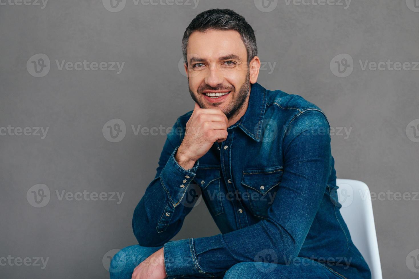 affascinante bello. fiducioso maturo uomo nel jeans camicia Tenere mano su mento e sorridente mentre seduta su il sedia contro grigio sfondo foto