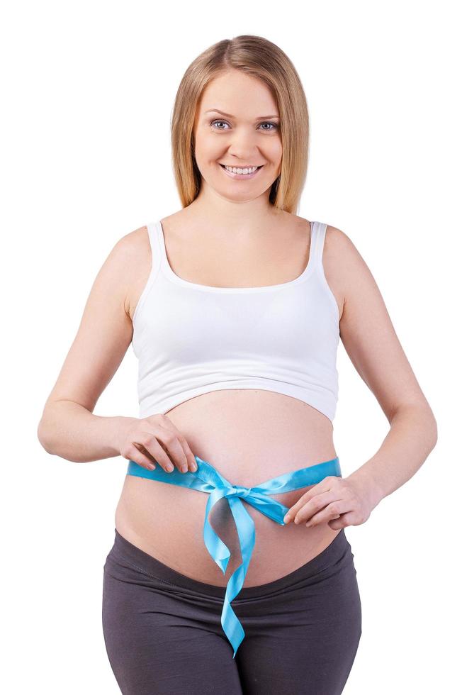 in attesa per un' poco ragazzo. contento incinta donna con blu nastro su sua pancia guardare a telecamera e sorridente mentre in piedi isolato su bianca foto