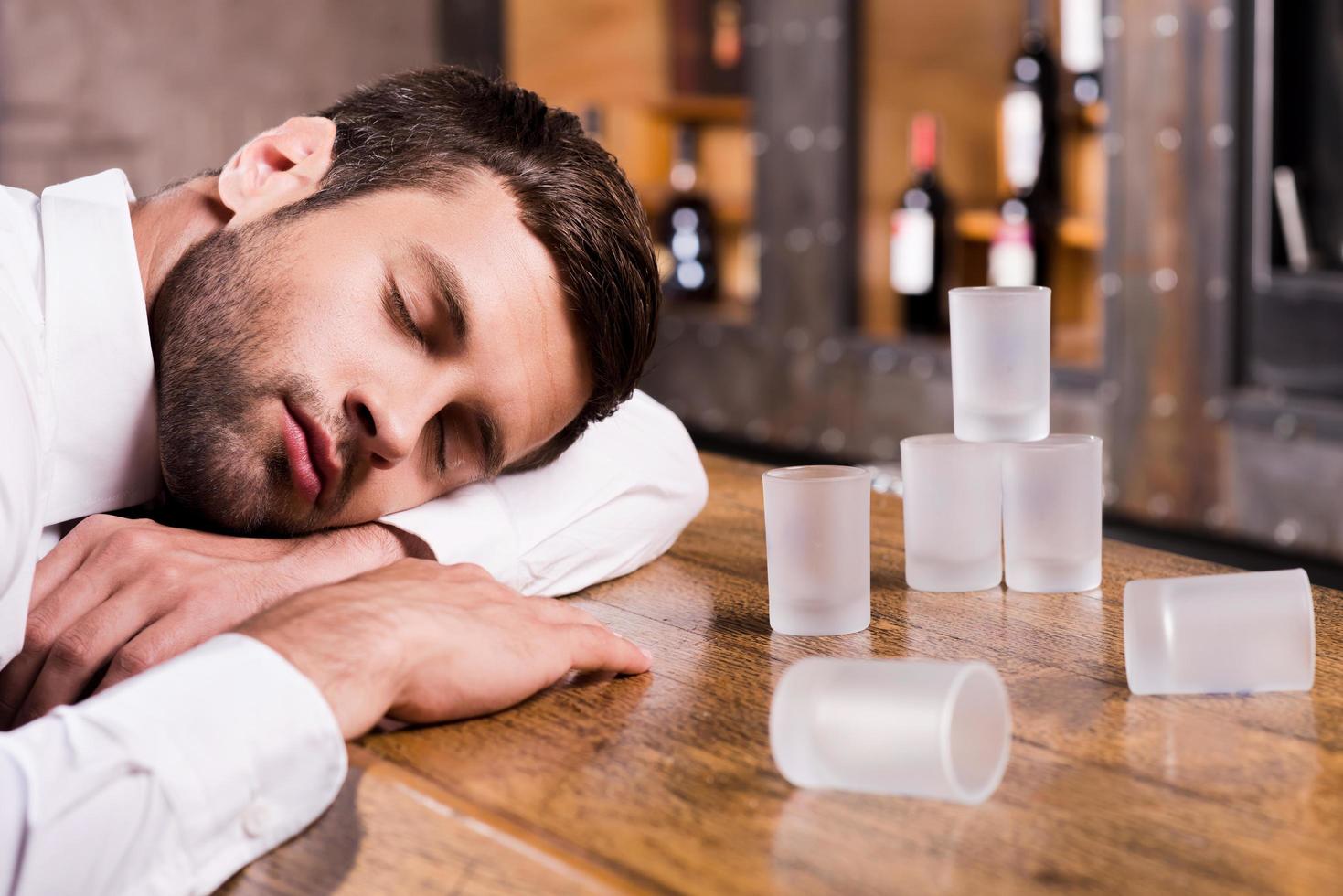 ubriaco ancora. ubriaco uomo nel bianca camicia pendente a il bar contatore e addormentato mentre vuoto bicchieri in piedi vicino lui foto