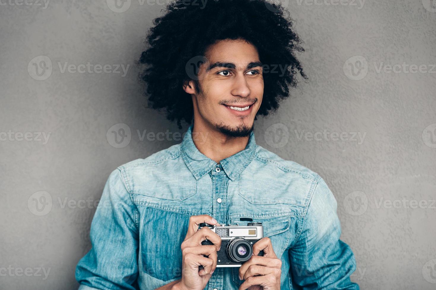 moderno fotografo. bello giovane africano uomo Tenere retrò styled telecamera e guardare lontano con Sorridi mentre in piedi contro grigio sfondo foto