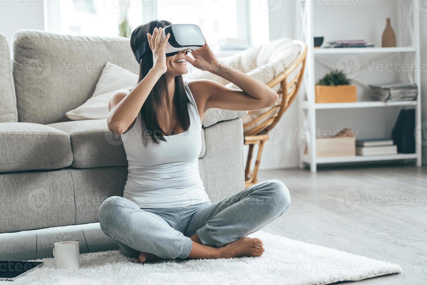 esso è così vero attraente giovane donna regolazione sua vr cuffia e sorridente mentre seduta su il tappeto a casa foto