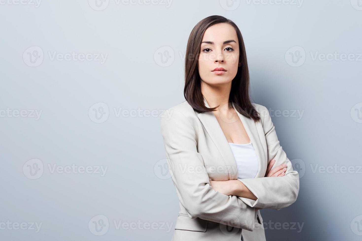 affascinante donna d'affari. fiducioso giovane donna d'affari conservazione braccia attraversato e guardare a telecamera mentre in piedi contro grigio sfondo foto