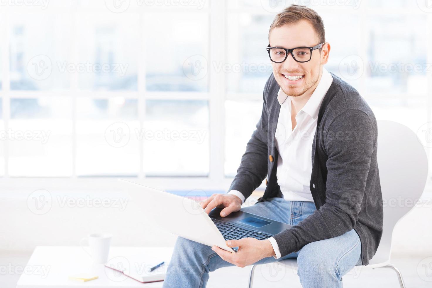 creativo esecutivo. bello giovane uomo nel bicchieri Lavorando su computer e sorridente a telecamera mentre seduta a il suo Lavorando posto foto