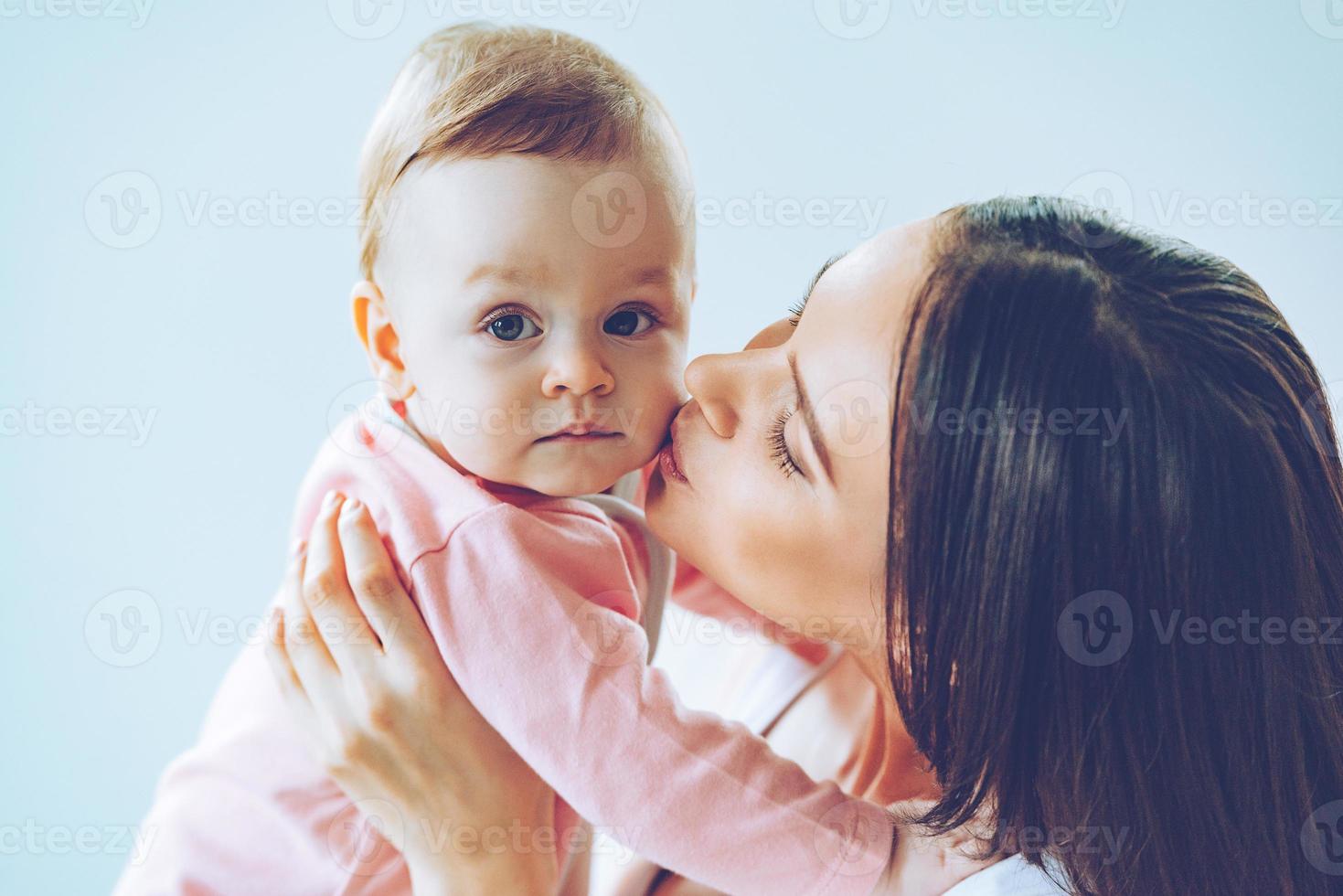 madre bacio. avvicinamento di bellissimo giovane donna Tenere bambino ragazza nel sua mani e baci sua con amore mentre seduta contro grigio sfondo foto