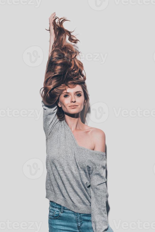 lungo e salutare capelli. attraente giovane donna allungamento sua capelli su e sorridente mentre in piedi contro grigio sfondo foto