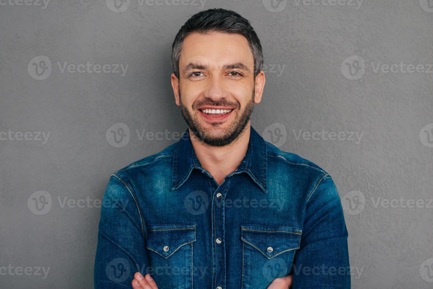 allegro bello. fiducioso maturo uomo nel jeans camicia conservazione braccia attraversato e sorridente mentre in piedi contro grigio sfondo foto