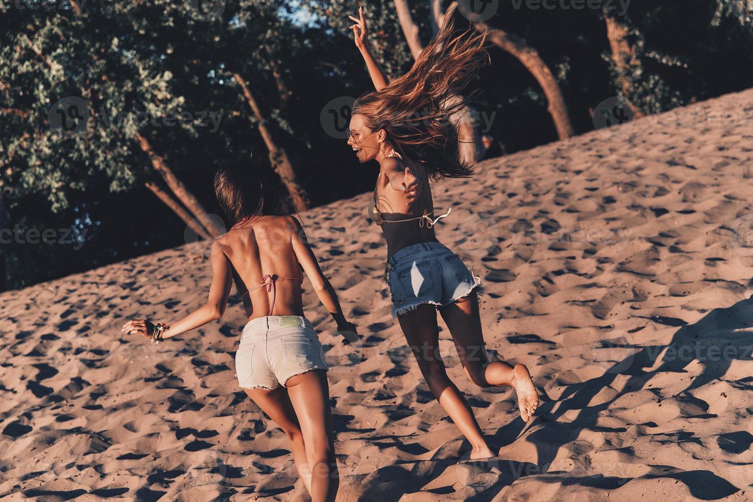 avendo grande tempo insieme. Due attraente giovane donne nel pantaloncini e costumi da bagno sorridente mentre in esecuzione su il spiaggia foto