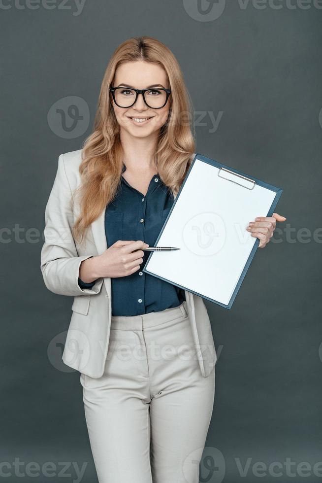 io bisogno il tuo firma Qui bellissima donna nel inteligente casuale indossare puntamento su vuoto carta e guardare a telecamera mentre in piedi contro grigio sfondo foto