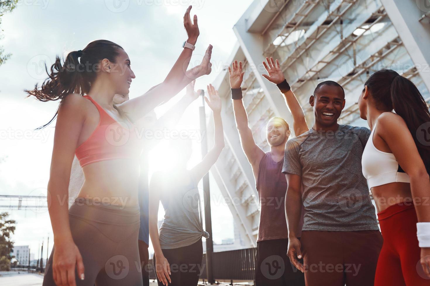 gruppo di giovane persone nel gli sport capi di abbigliamento dando ogni altro alto cinque e sorridente mentre esercizio all'aperto foto