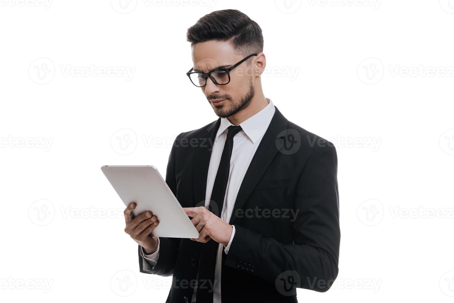 l'esame il suo digitale tavoletta. grave giovane uomo nel pieno completo da uomo Lavorando su il suo digitale tavoletta mentre in piedi contro bianca sfondo foto