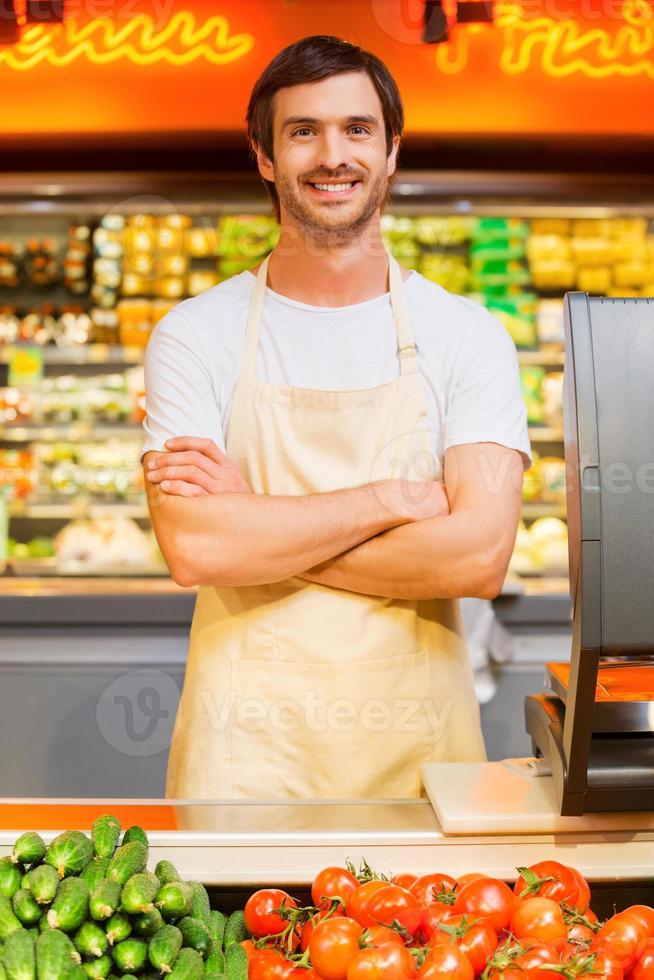 Come Maggio io Aiuto voi bello giovane cassiere conservazione braccia attraversato e sorridente mentre in piedi a supermercato check-out foto