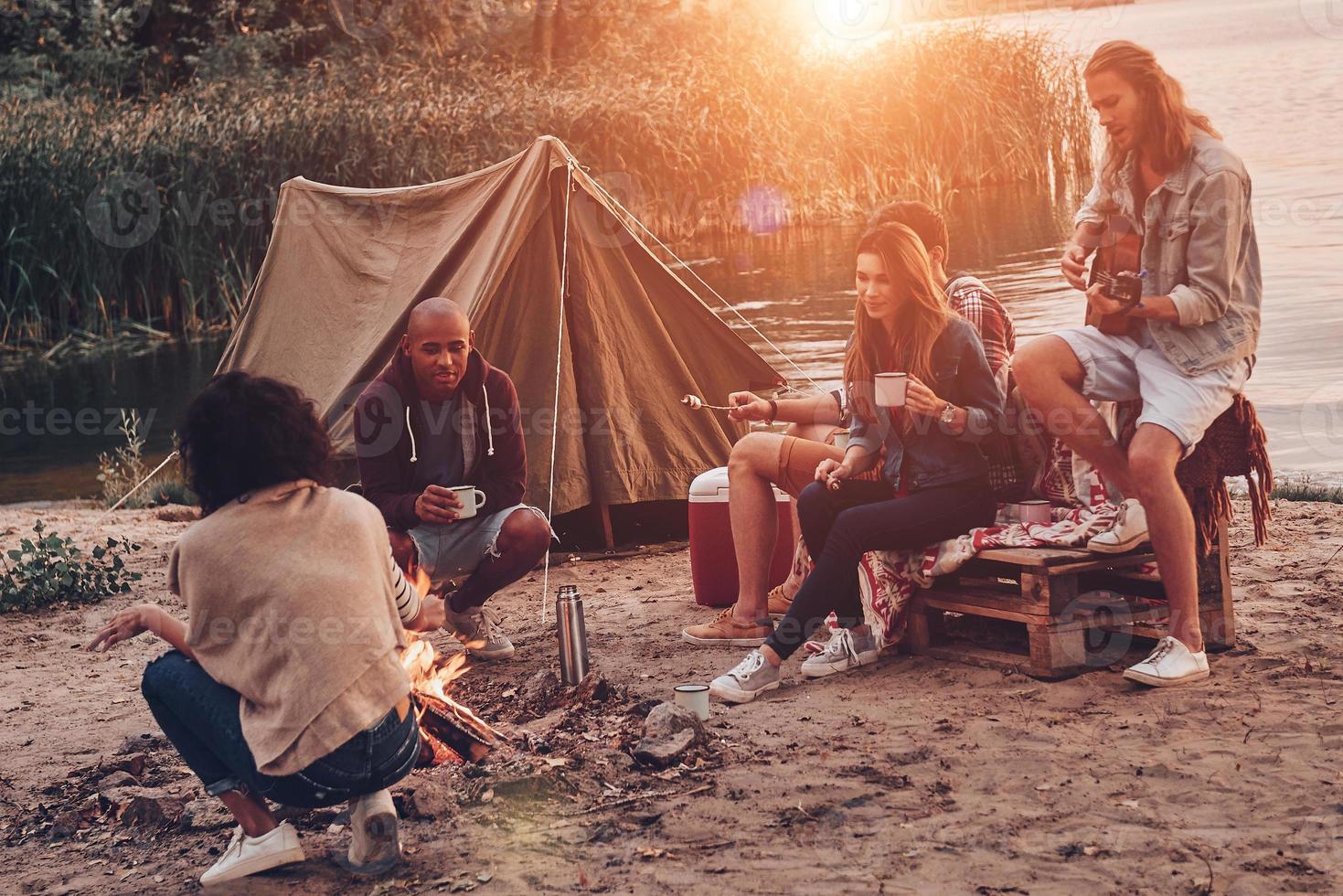 momenti di la gioia. gruppo di giovane persone nel casuale indossare sorridente mentre godendo spiaggia festa vicino il fuoco di bivacco foto